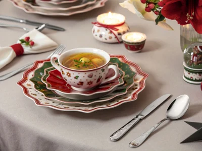 Toys Delight traditional christmas tablesetting with green red and white plates A festive table setting with a Toys Delight bowl of soup in a decorated cup, surrounded by candles and Christmas-themed decor.