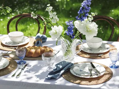 Ein Gartentisch ist mit Blumenporzellan „Alt Luxemburg“ von Villeroy & Boch, Tischsets aus Korbgeflecht, blau getöntem Glasgeschirr, einer Kristallkanne mit blauen und weißen Blumen und einem Brotlaib gedeckt.