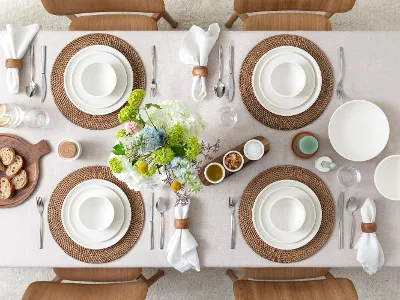An elegantly set dining table with woven placemats, white plates, napkin rings, and flowers as table decor. Bread on a wooden tray and small bowls are also on the table.