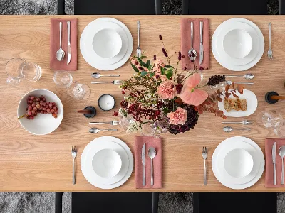 Apparecchiatura floreale e uva Un tavolo da pranzo in legno con piatti bianchi, ciotole, posate su tovaglioli rosa, due candele, una ciotola con uva e una composizione floreale.