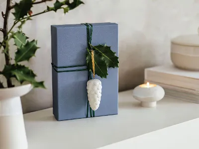 Blaue Geschenkbox geschmückt mit Winter Glow Ornament Blaue Geschenkbox, geschmückt mit Winter Glow Ornament, neben einer brennenden Kerze und Büchern auf einem Tisch.