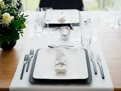 Une table de salle à manger avec des assiettes carrées Manufacture Une table de salle à manger avec des assiettes carrées Manufacture, une serviette roulée avec un rond de serviette argenté, des couverts de table et plusieurs verres.