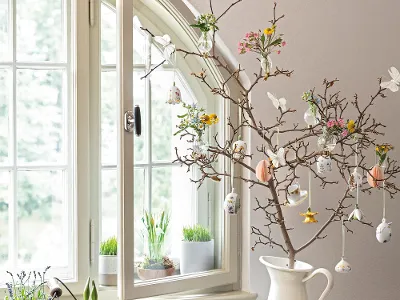 Mi Osterornamenten von Villeroy & Boch geschmückter Baumzweig auf der Fensterbank Eine frühlingshafte Inneneinrichtung mit einem dekorativen, mit Osterornamenten von Villeroy & Boch geschmückten Baumzweig auf der Fensterbank, begleitet von Pflanzen und einem weißen Krug auf einem Tablett.