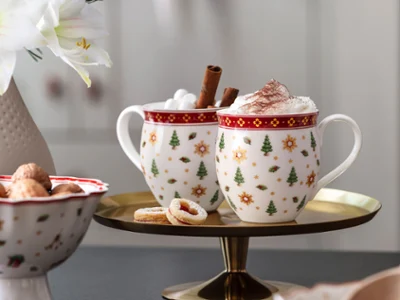 Two festive Toy's Delight cups with whipped cream and cinnamon sticks on a tray, with biscuits and a flower vase next to them.