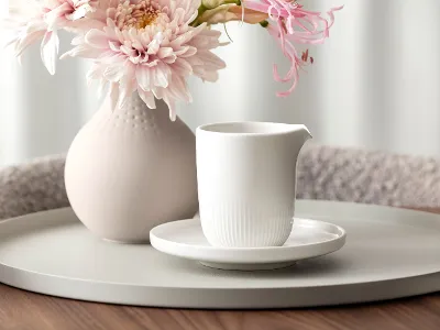 Afina milk jug staged A white ceramic pitcher on a saucer stands on a round tray next to a vase with pink flowers on a wooden table.