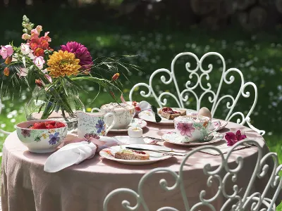 Ein Mariefleur-Gartentisch-Set von Villeroy & Boch besteht aus Porzellan mit Blumenmuster, einer Vase voller bunter Blumen und Tellern, die mit Desserts und Erdbeeren geschmückt sind. Verzierte weiße Stühle umgeben den Tisch vor einem Hintergrund aus üppigem grünem Gras.