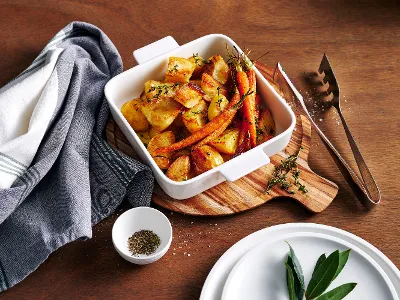 aus-clevercooking-indoor-food-nopeople-squaredish3245 The 21cm Clever Cooking square baking dish, filled with freshly roasted potatoes and carrots, sits atop a circular wooden board on a rustic wooden table. Complemented by silver serving tools and a blue and white linen napkin, the dish is ready to serve alongside white plates.