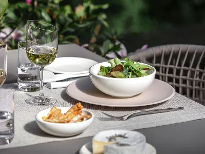 Un plat creux NewMoon blanc avec de la salade sur une assiette plate beige Un plat creux NewMoon de Villeroy & Boch rempli de salade et des verres à vin.