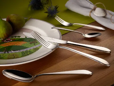 Sereno Besteckset auf Design Naif Teller angeordnet auf einem Holztisch. Ein weißer Teller mit Landschaftsmotiv, zwei Feigen und ein Sereno Besteckset sind auf einem Holztisch angeordnet.