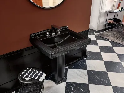 Schwarzes Waschbecken mit schwarzem Wasserhahn aus der Kollektion Hommage Ein schwarzes Hommage Waschbecken mit einem modernen Wasserhahn vor einer roten Wand, ein weiß-schwarz karierter Boden und ein kleiner runder Tisch mit einem gefalteten Handtuch.