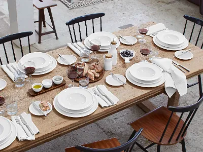 Artesano Geschirr auf hölzernem Esstisch Ein hölzerner Esstisch für sechs Personen mit weißem Geschirr, Rotweingläsern, Servietten, Brot, Trauben, Butter, Salz, Pfeffer, Öl und Wassergläsern. An jedem Gedeck sind Teller und Besteck aufgestellt.