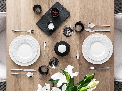 De la vaisselle Manufacture avec des couverts de table Piemont sur une table moderne dressée pour 2 personnes Une table de salle à manger joliment dressée avec deux assiettes blanches Manufacture, des couverts de table Piemont, des verres, une bougie, un plateau noir avec des aliments et un vase avec des lys blancs sur une surface en bois.