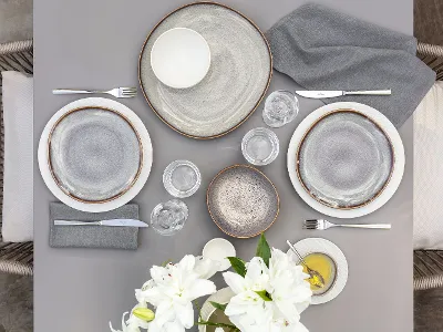 Top view of a dining table with two place settings, consisting of beige Lave plates and white Manufacture plates, cutlery, glasses and a vase of white flowers.