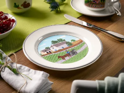 On the table is a decorative plate from Villeroy & Boch Design Naif, which depicts a picturesque landscape with a house, trees, and a horse-drawn carriage. The accompanying items include a knife, a cup, a bowl of red berries, and a green cloth.