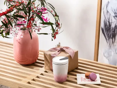 Un vase Perlemor rose avec des fleurs et une tasse Coffee ToGo de Villeroy & Boch sont posés sur un banc en bois. Un vase Perlemor rose avec des fleurs, une boîte cadeau avec un ruban et un gobelet Coffee ToGo de Villeroy & Boch sont posés sur un banc en bois.