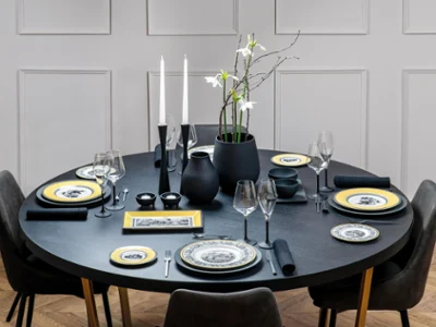 Full table set with Manufacture Rock and Audun Elegant round dining table with black tableware, yellow-rimmed plates, white flowers in a vase, and lit candles, set in a modern room.