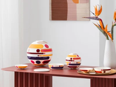 Colorful, multi-piece tableware sets from la petite boule miami, circular design objects in large and small sizes, stand as decoration next to a vase with abstract patterns and orange flowers on a red table.