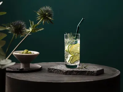 Glas von La Divina von Villeroy & Boch stilvoll serviert Ein La Divina Glas mit Gurkenscheiben und Rosmarin, auf einer Steinfläche mit grünem Hintergrund. Ein Artesano Glasstrohhalm steckt im Getränk und daneben steht eine Schale mit Oliven.