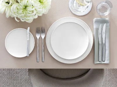 Tischgedeck mit Afina Geschirr und Piemont Besteck Anspruchsvolles Tischarrangement mit der Piemont-Kollektion von Villeroy & Boch, einschließlich eines weißen Esstellers, eines Salattellers, eines Bestecksets, Glaswaren und eines floralen Tafelaufsatzes auf einer beigen Tischdecke.
