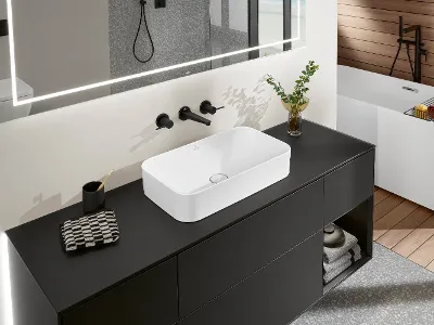 weißes Finion-Waschbecken auf schwarzer Arbeitsplatte mit schwarzer Armatur Ein modernes Badezimmer mit einem rechteckigen Finion-Waschbecken auf einer schwarzen Arbeitsplatte, einem Spiegel darüber, schwarzen Armaturen und einer Topfpflanze. Im Hintergrund ist eine Badewanne zu sehen.