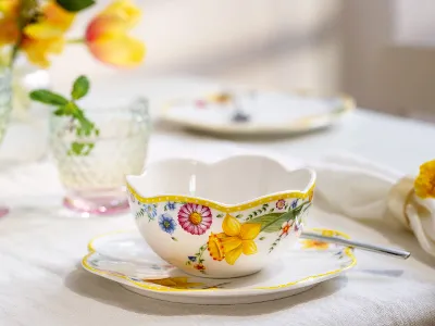Die Teetasse und Untertasse „Spring Awakening“ mit Löffel von Villeroy & Boch. Die Teetasse und Untertasse „Spring Awakening“ mit Löffel von Villeroy & Boch stehen auf dem Tisch, neben Kristallgläsern und sanft verschwommenen Blumen im Hintergrund.