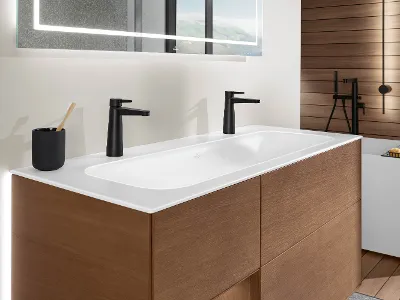 Zwei Finion-Waschbecken mit schwarzen Armaturen Modernes Waschbecken mit weißer Arbeitsplatte, zwei schwarzen Wasserhähnen, einem schwarzen Zahnbürstenhalter mit Zahnbürste und darunter Holzschränken. An der Wand darüber hängt ein großer Spiegel mit integrierter Beleuchtung.