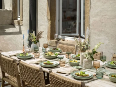 A rustic outdoor dining table is set with Perlemor plates of salad A rustic outdoor dining table is set with Perlemor plates of salad, wine glasses, and floral centerpieces by a stone wall.