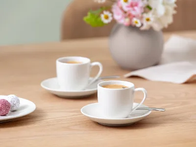 Due tazze ForMe bianche di Villeroy & Boch su piattini con cucchiai su un tavolo di legno. Due tazze ForMe bianche di Villeroy & Boch su piattini con cucchiai su un tavolo di legno. Sullo sfondo un piatto con tartufi colorati e un vaso con fiori bianchi e rosa.