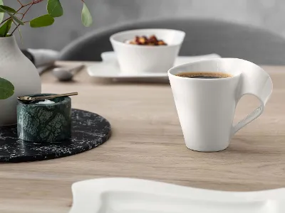 Un mug NewWave avec un vase de fleurs Une tasse à café blanche moderne avec un design d’anse unique, placée à côté d’un petit récipient et d’un vase avec des feuilles vertes sur une table en bois.