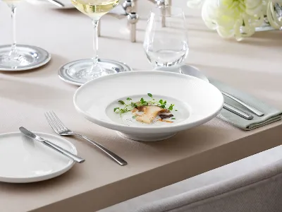 Une assiette creuse Afina blanche remplie de soupe Une assiette creuse Afina de Villeroy & Boch contenant une soupe de cèpes à la crème, à côté d’un verre à vin blanc ainsi que des couverts de table Piemont.