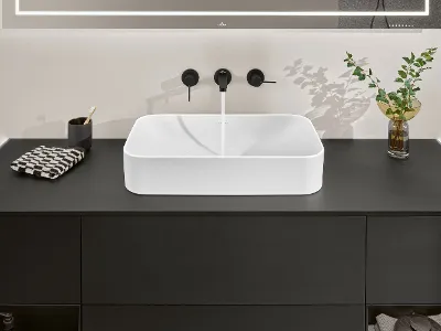 Umywalka Finion z czarną baterią i blatem Stylowa umywalka Villeroy & Boch z białą prostokątną misą, czarnym blatem, czarnym kranem, składanym ręcznikiem, uchwytem na szczoteczkę do zębów i wazonem