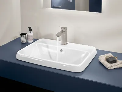 Weißes Architectura Waschbecken mit silbernem Wasserhahn Eine weißes rechteckiges Architectura Waschbecken mit fließendem Wasser, einem silbernen Wasserhahn, einem beigefarbenen Handtuch und einem Behälter mit Flüssigseife auf einem blauen Regal in einem Badezimmer.