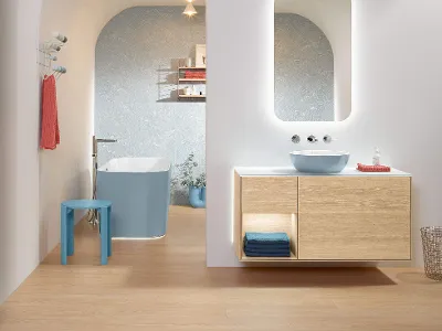 Hellblaues Artis Waschbecken in hellem Badezimmer Modernes Badezimmer-Interieur mit hellblauem Artis Waschbecken, freistehender Finion Badewanne und Holzwaschtisch sowie einem runden, beleuchteten More to See Spiegel