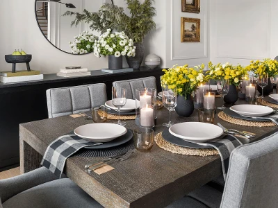 Modern dining room with a dark wood table set for six, featuring gray chairs, yellow flowers, candles, and a large round mirror on the wall.