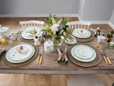 Dezent gedeckter Ostertisch mit Colourful Spring Geschirr Ein Tafelset „Colourful Spring“ für vier Personen von Villeroy & Boch besteht aus Porzellan mit Blumenmuster, Besteck mit gelben Griffen, gewebten Tischsets und einem Blumengesteck. Zu jedem Gedeck gehört ein Ei in einer dekorativen Tasse.
