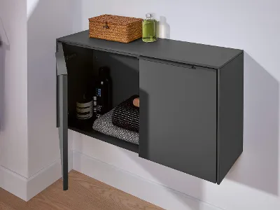 Ein anthrazit-farbenes Sideboard mit geöffneter Tür, durch die man Badtextilien und Accessoires sehen kann. Auf dem Sideboard steht eine kleine Box und ein Glas mit grüner Flüssigseife.
