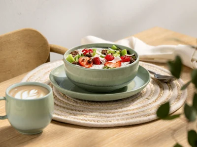 A green Perlemor bowl filled with yoghurt and a Perlemor coffee cup A bowl of yoghurt with fruit and muesli next to a cup of coffee on a table with a woven placemat.