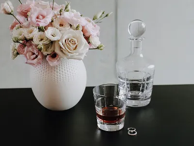 Un vaso Manufacture con fiori bianchi e rosa, un bicchiere di whisky, una caraffa per whisky. Un vaso con fiori bianchi e rosa, un bicchiere di whisky, una caraffa e due anelli su un tavolo nero.
