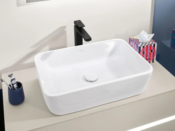 Biała umywalka Architectura z czarną baterią Beżowy blat z prostokątną białą umywalką z kolekcji Architectura z elegancką czarną baterią. Obok stoją różne akcesoria kąpielowe.