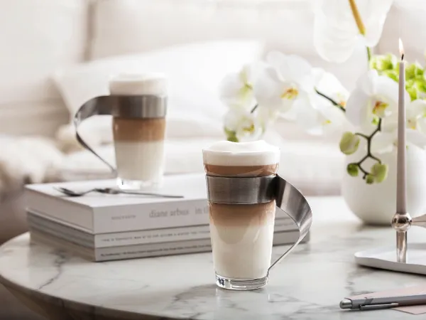 Zwei Kaffeegetränke in NewWave Latte Macchiato Gläsern mit eleganten Metallgriffen. Zwei Kaffeegetränke in NewWave Latte Macchiato Gläsern von Villeroy & Boch mit eleganten Metallgriffen werden auf einem Marmortisch platziert, umgeben von Büchern, Orchideen und einem Kerzenständer.