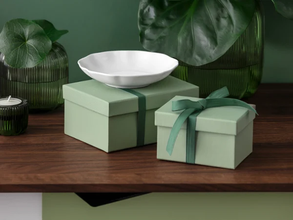 Zwei grüne Geschenkschachteln stehen auf einer Holzfläche mit einer weißen Pura Schale Zwei grüne Geschenkschachteln, eine davon mit einer Schleife, stehen auf einer Holzfläche mit einer weißen Pura Schale und grünen Glasdekorationen.