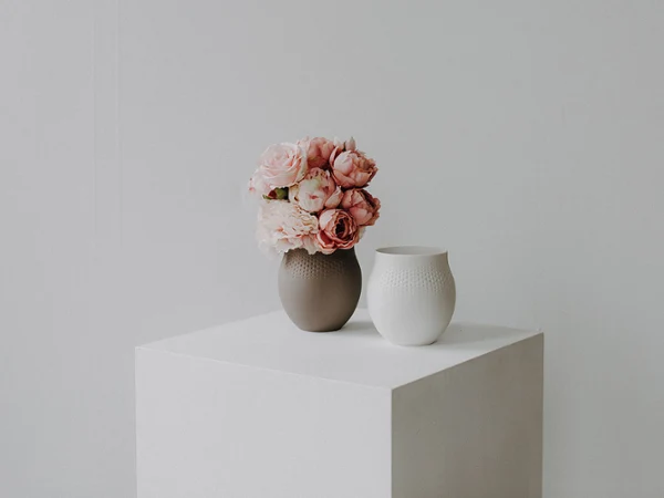 Brązowy wazon z różowymi kwiatami i biały ceramiczny wazon stoją na białej kostce na jednolitym jasnym tle.
