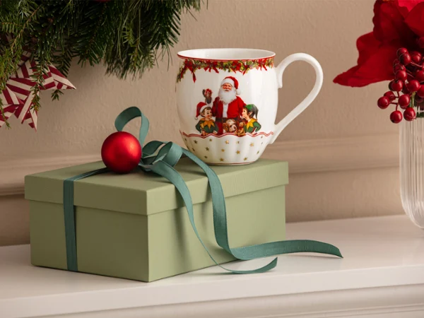 A green gift box with a bow, a red Christmas ornament, and a mug with a Santa Claus motif from Villeroy & Boch in front of a white background, with Christmas decorations visible in the background.