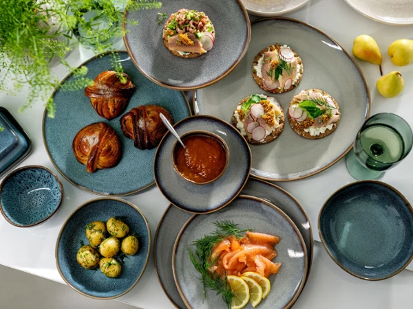 Eine Auswahl an Gourmetgerichten, darunter Räucherlachs, belegte Brötchen und Gebäck, serviert auf stilvollen Lave-Tellern von Villeroy & Boch auf einem weißen Tisch.