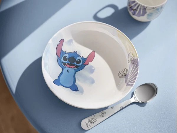 Un cuenco blanco de Disney Stitch, una cuchara a juego y una taza sobre una mesa. Un cuenco blanco de Disney Stitch con un motivo de dibujos animados, una cuchara a juego y una taza sobre una mesa azul a la luz del sol.