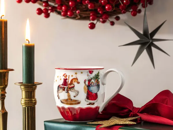 Le mug Annual Christmas Edition 2024 dans une scène de Noël Une scène de Noël festive montre la coupe annuelle décorée Annual Christmas 2024 avec le Père Noël, deux bougies allumées dans des supports en laiton, un ruban rouge, un ornement en forme d’étoile et une couronne de baies rouges en arrière-plan.