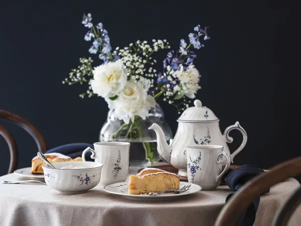 Une table ronde dressée avec du thé Vieux Luxembourg, des tasses à thé, des parts de gâteau et un vase de fleurs blanches sur fond sombre.