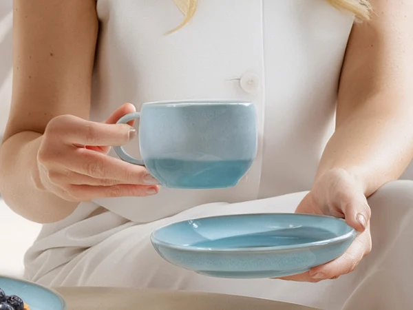 Una taza de cerámica azul nacarada Aqua y un platillo a juego. Una persona ligeramente vestida sostiene una taza de cerámica azul Perlemor Aqua en una mano y un platillo a juego en la otra.