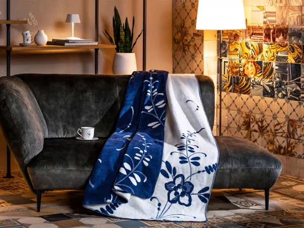 Koce Vieux Luxembourg Ciemna sofa z niebiesko-białym wzorzystym kocem nad nią, lampa obok i półka z przedmiotami dekoracyjnymi.