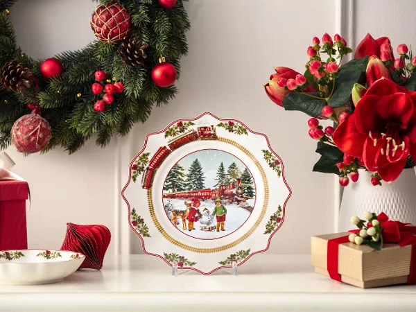 A decorative Christmas plate featuring a winter scene is displayed on a mantelpiece adorned with Christmas garlands, flowers, and wrapped presents.